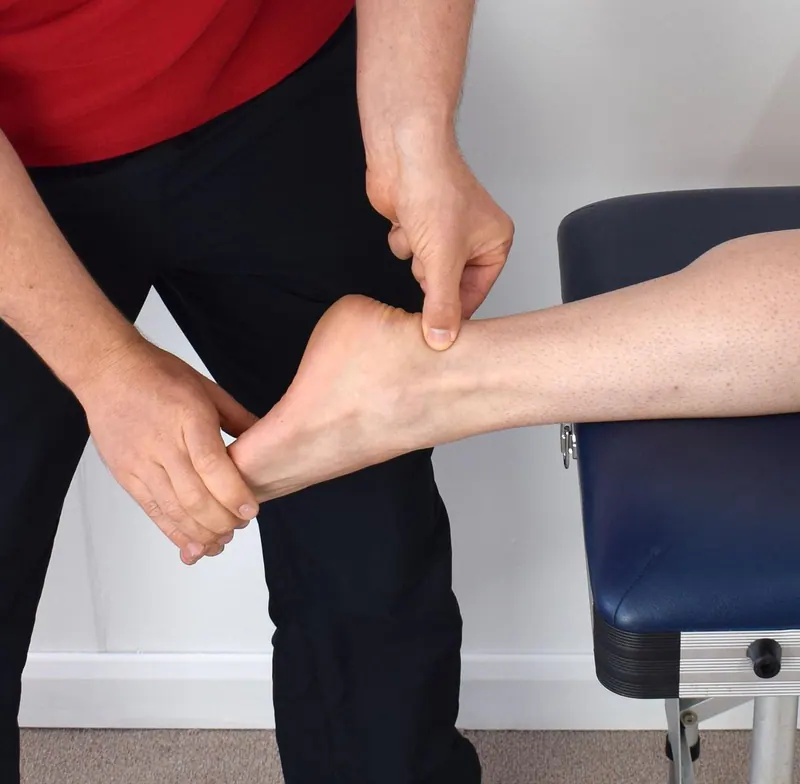 Hände eines Therapeuten stabilisieren Ferse und Sprunggelenk eines Beins auf einer Therapieliege